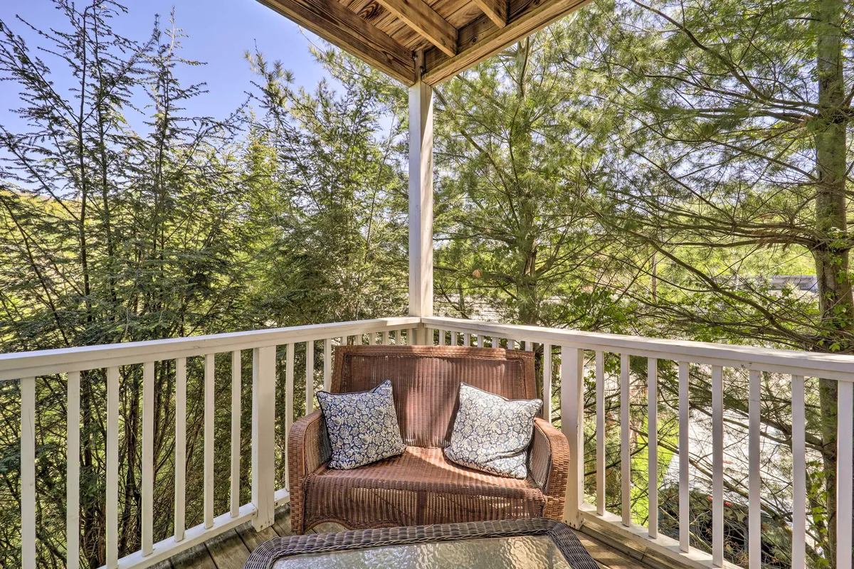 A portion of the main covered deck featuring a cozy wicker couch adorned with decorative pillows, set against a sturdy wooden railing, with lush green foliage visible beyond. The tranquil scene invites relaxation and offers a direct connection to the surrounding nature, ideal for morning coffee, reading, or socializing.