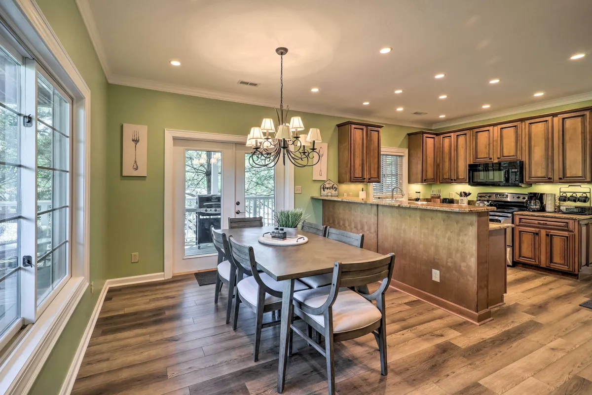 A view of a cheerful dining room featuring a large wooden table, an elegant chandelier overhead, and a kitchen counter in the background. The room is styled for comfort and group gatherings, suggesting lively meals and celebrations.