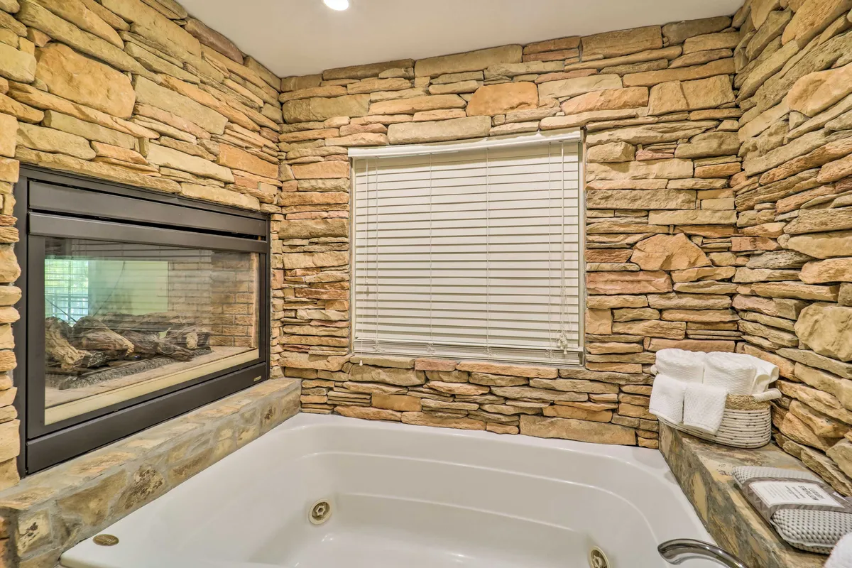 A luxuriously appointed bathroom with a stone wall, fireplace, jacuzzi tub, and a window letting in soft light. Towels are neatly arranged in a basket, creating a spa-like atmosphere.