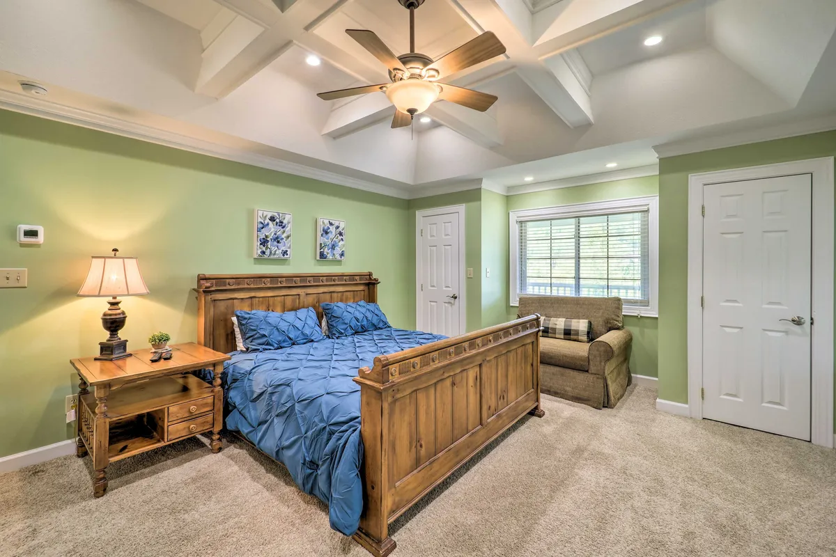 A cozy king bedroom with a wooden bed frame and blue bedding, complemented by a ceiling fan, plush chair, and decorative wall art. The room is filled with soft natural light, evoking a peaceful and restful mood.