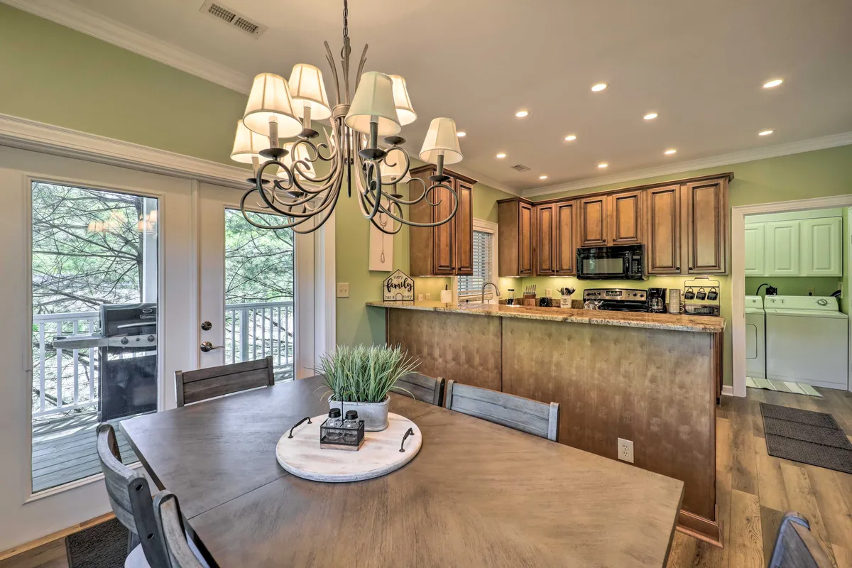 A warmly lit dining area with a prominent chandelier, a spacious dining table ready for gatherings, open kitchen cabinetry, and a window with scenic views. The space radiates a welcoming, festive atmosphere perfect for group meals and entertaining.